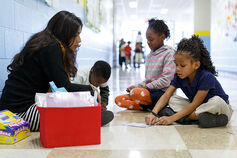 A student mentor works with elementary students in a school hallway.